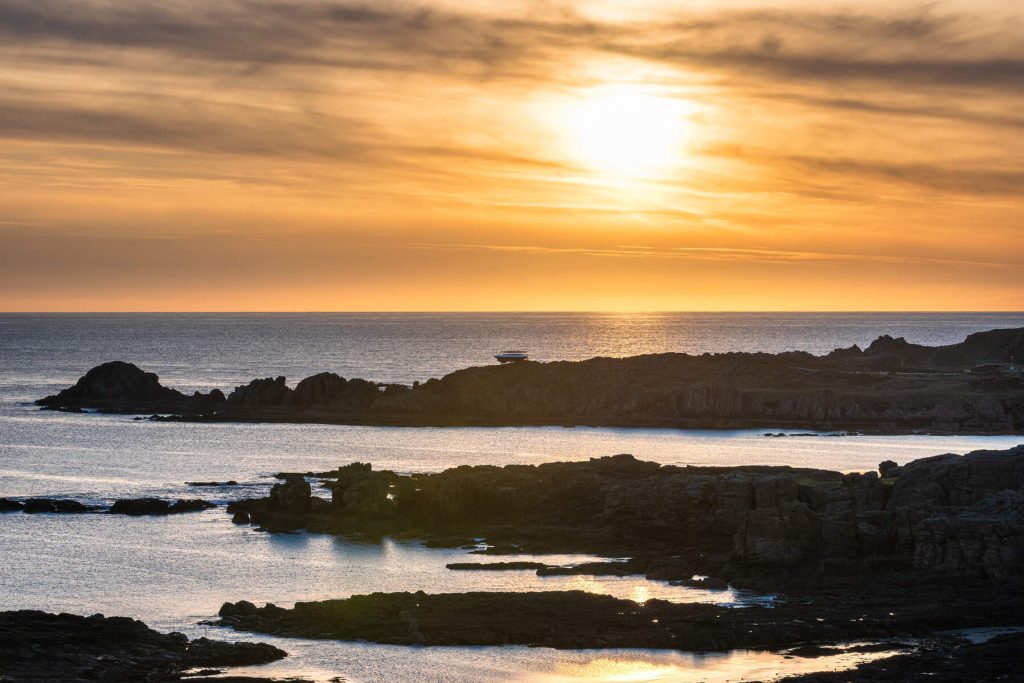 Golden sunset over a rocky coastline, with the sun low in the sky casting warm reflections on calm ocean water and silhouetted rock formations in the foreground.
