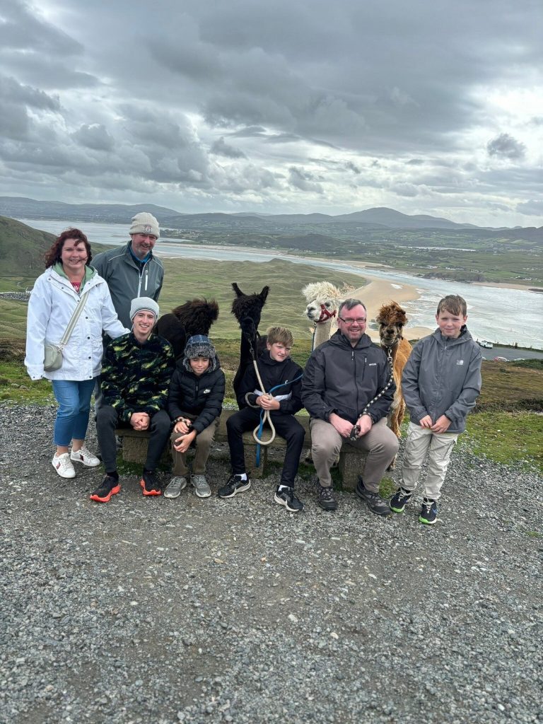 A group of seven people—four adults and three children—pose outdoors on a hilltop with three alpacas. The sea, sandy shoreline, fields, and mountains are visible under a cloudy sky in the background.