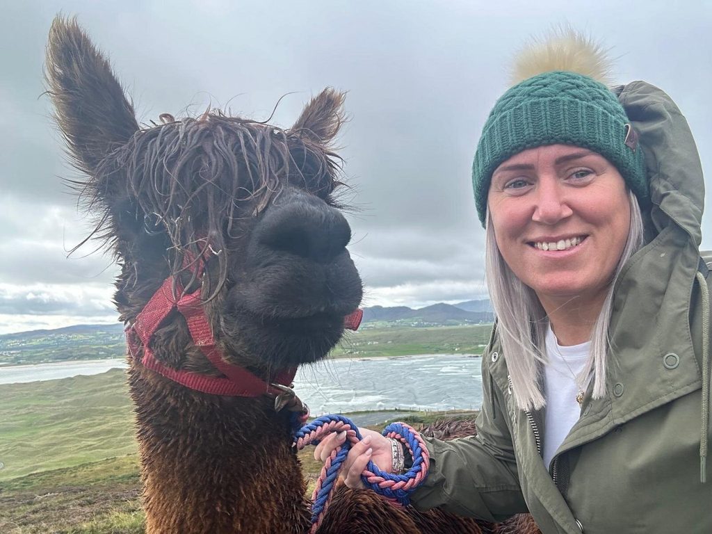 A woman in a green beanie and jacket smiles while holding a leash next to a brown alpaca with a red halter. They are outdoors with hills, water, and cloudy skies in the background.