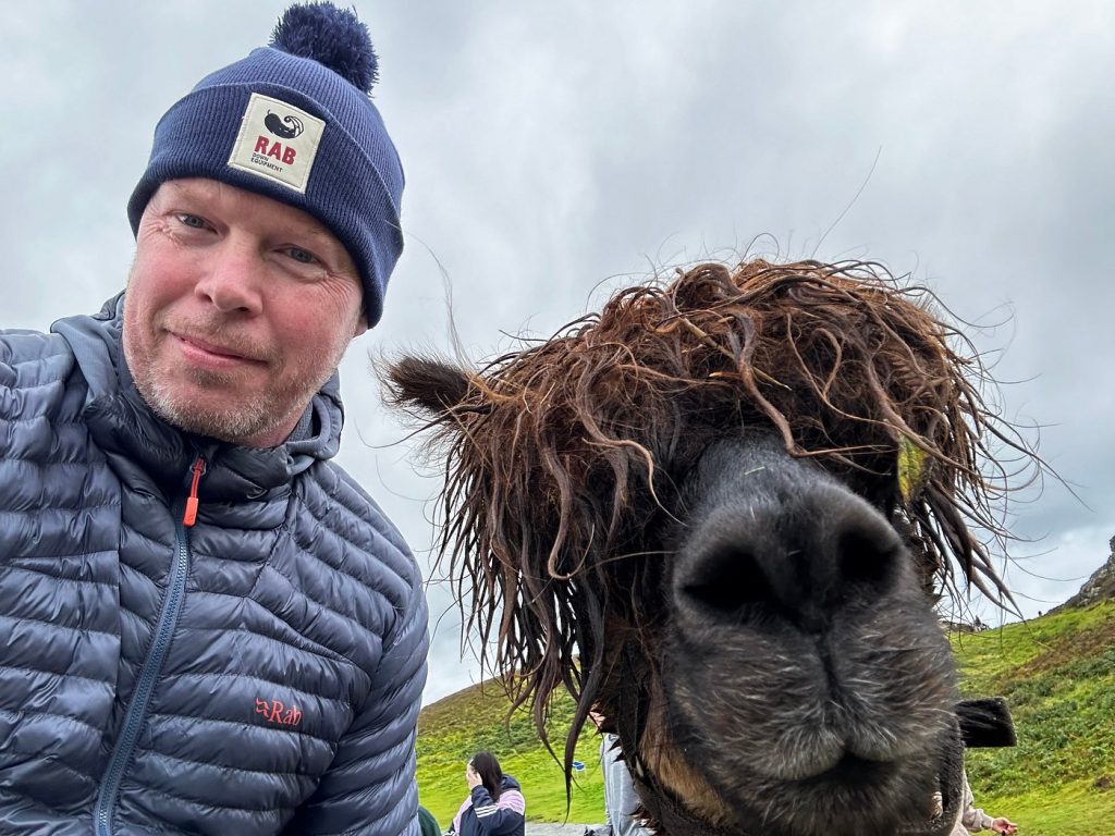 A man in a blue beanie and puffer jacket takes a close-up selfie with a black alpaca with shaggy hair. The background shows grassy hills and people in outdoor clothing under a cloudy sky.