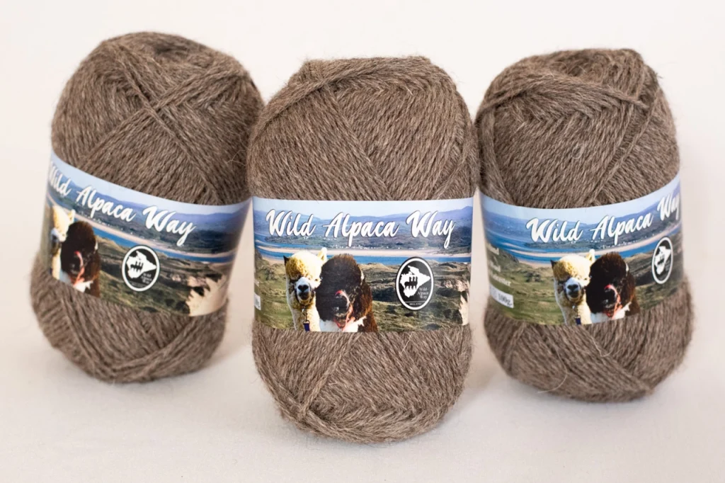 Three brown skeins of yarn labeled "Wild Alpaca Way" with an image of alpacas and a scenic landscape on the packaging are arranged side by side on a white background.